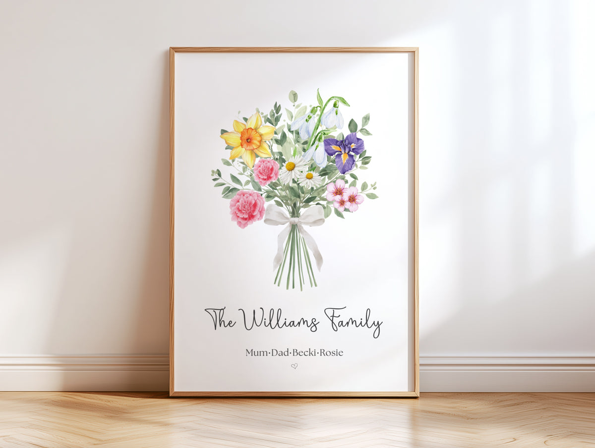 A framed personalised watercolor birth flower family print leaning against a white wall on a light wood floor. The print features a bouquet of yellow, pink, purple, and white flowers with green leaves, personalized with the text 'The Williams Family' and 'Mum-Dad-Beckie-Rosie'.