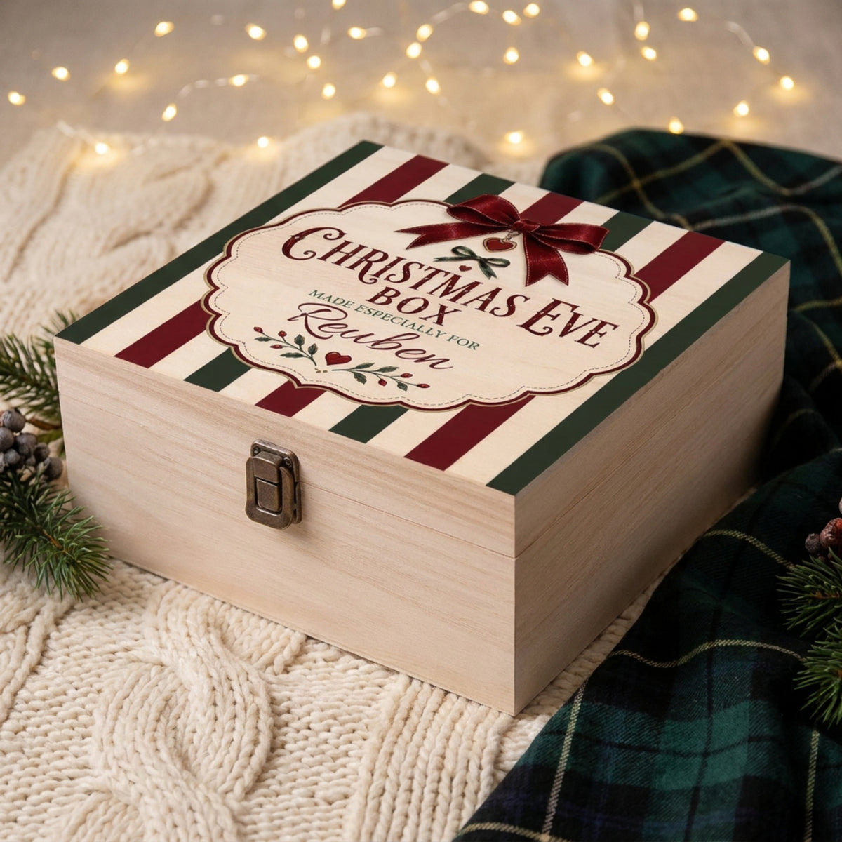 A personalized wooden Christmas Eve box with a striped lid in cream, burgundy, and green, heritage design. featuring a custom printed design that reads 'Christmas Eve Box Made Especially for Ruben' with a bow illustration. The box is styled on a cream cable-knit blanket with a green plaid scarf and warm fairy lights in the background.